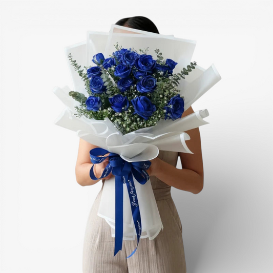 The Flowercart Sapphire Devotion Bouquet: 16 vibrant blue spray roses with baby's breath and eucalyptus, wrapped in white paper and tied with a blue ribbon.