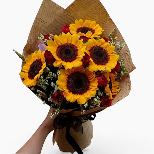 The Flowercart's Sunny Blooms bouquet features 6 golden sunflowers and 12 red roses with baby's breath, wrapped in brown paper.