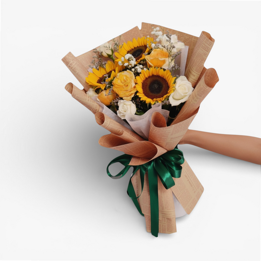 The Flowercart Sunny Serenade bouquet features sunflowers, yellow roses, white roses, and baby's breath, wrapped in brown paper with a green ribbon.