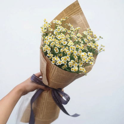 Bouquet of white flowers wrapped in brown paper with a purple ribbon, held against a light gray background.