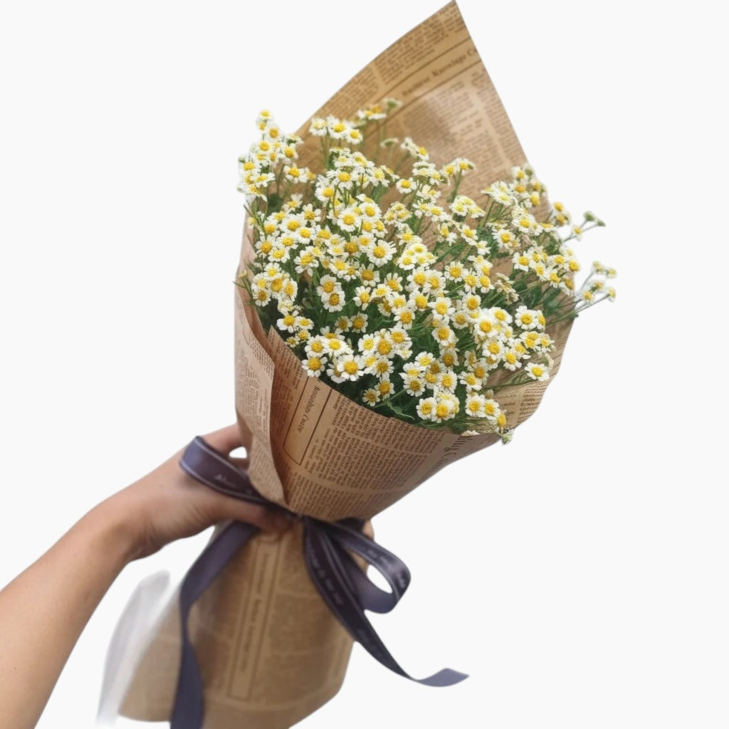 Bouquet of white flowers wrapped in brown paper with a purple ribbon, held against a light gray background.