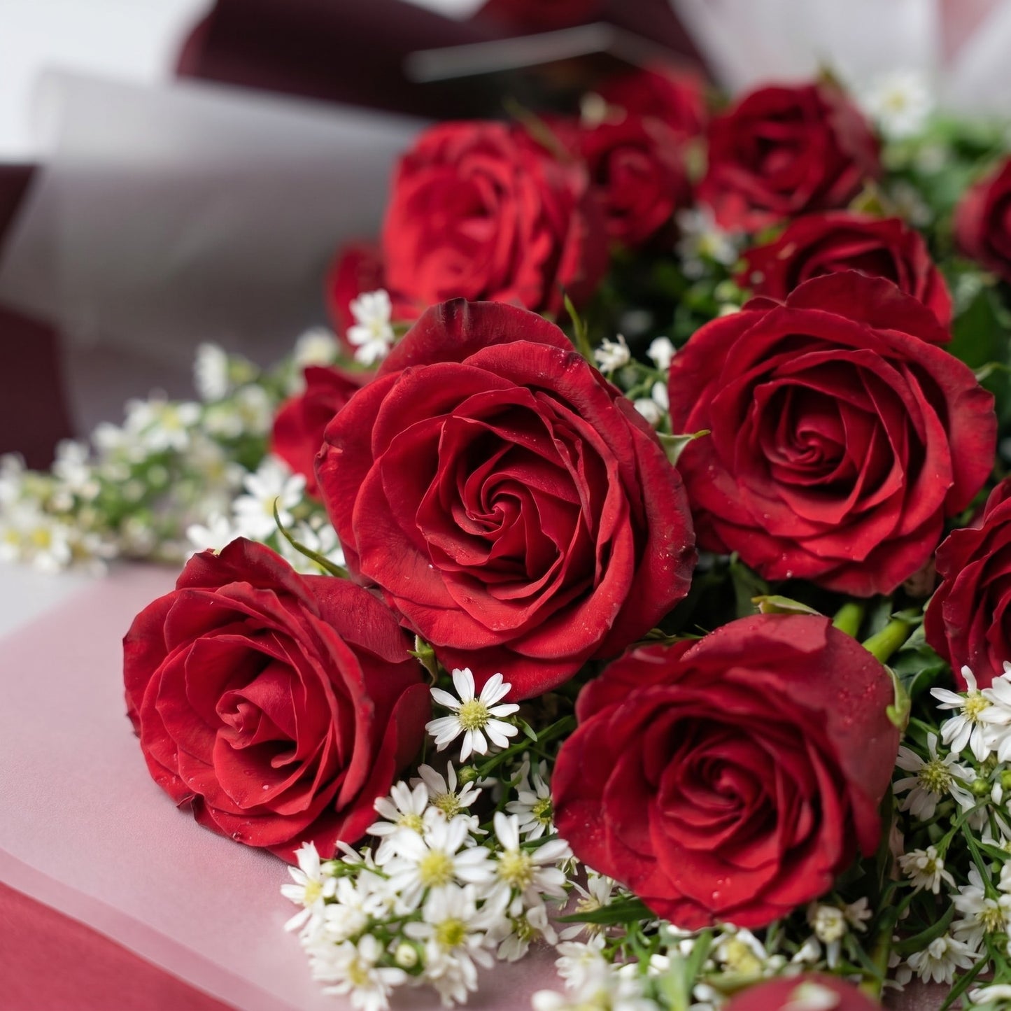 The Flowercart Crimson Whisper bouquet featuring 24 premium red roses and white filler flowers with a red ribbon.