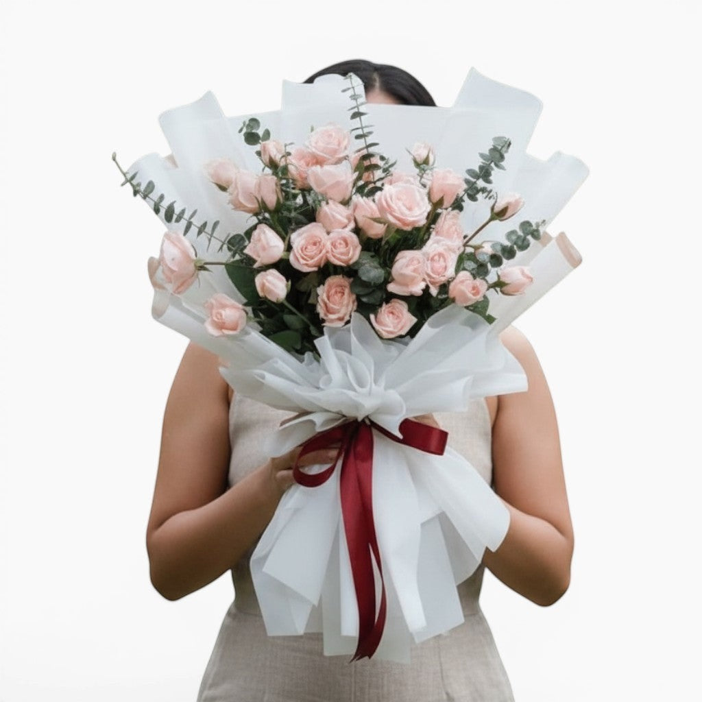 The Flowercart Lovely Spark bouquet featuring 24 baby pink roses and eucalyptus wrapped in white paper with a red ribbon