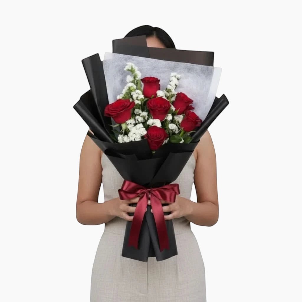 The Flowercart Hot Romance bouquet featuring six red roses and white statice wrapped in elegant black paper.