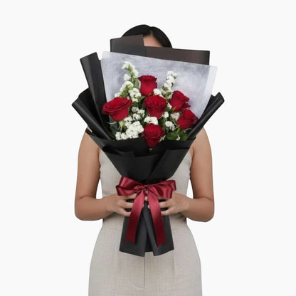 The Flowercart Hot Romance bouquet featuring six red roses and white statice wrapped in elegant black paper.