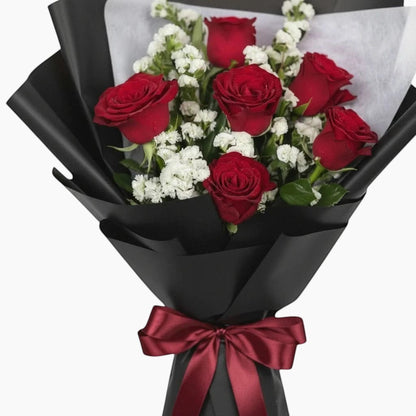 The Flowercart Hot Romance bouquet featuring six red roses and white statice wrapped in elegant black paper with a bow.