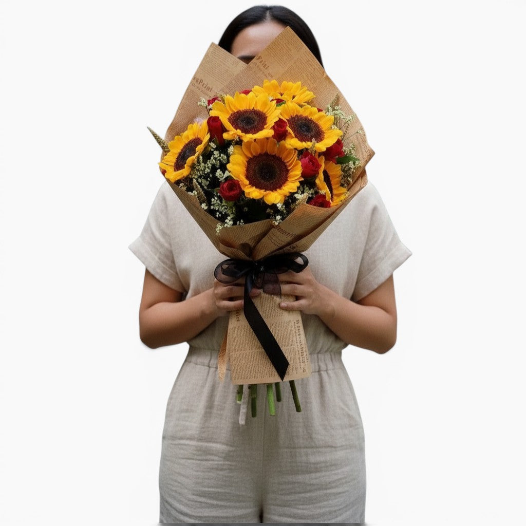 The Flowercart Sunny Blooms bouquet featuring sunflowers and red roses wrapped in brown paper held by a person.