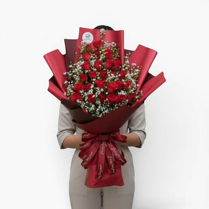 The Flowercart Valley Scent bouquet of 24 red roses and baby's breath wrapped in red paper with a decorative ribbon.