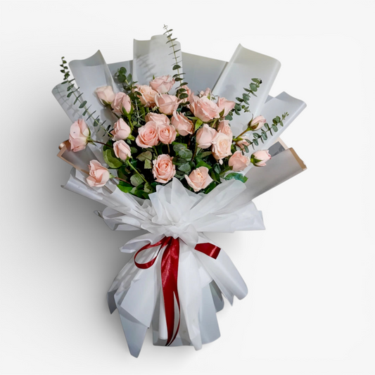 The Flowercart Lovely Spark bouquet: 24 baby pink roses with eucalyptus, wrapped in white paper and tied with a red ribbon.