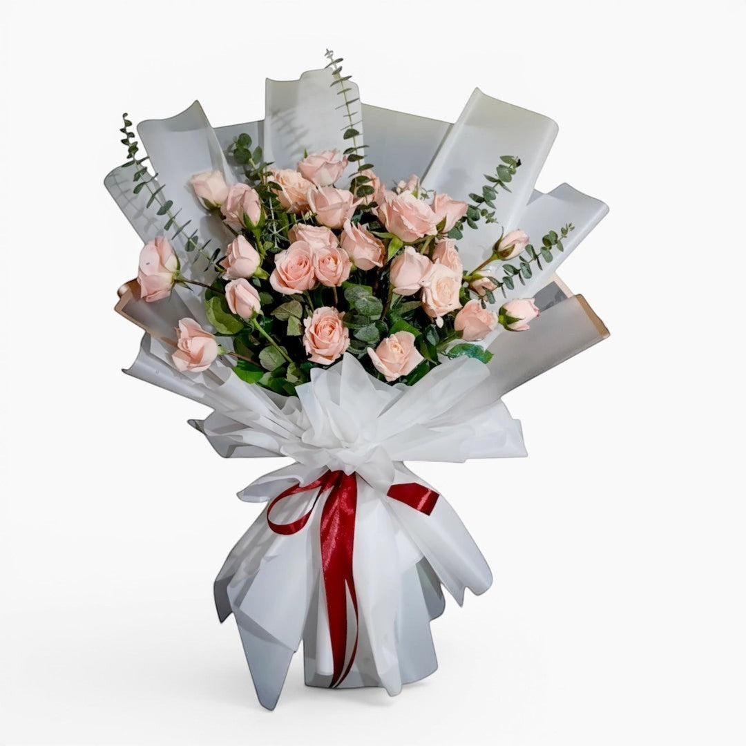 The Flowercart Lovely Spark bouquet: 24 baby pink roses with eucalyptus, wrapped in white paper and tied with a red ribbon.