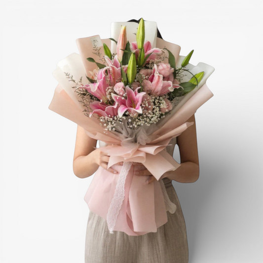 The Flowercart's Majestic Blush bouquet features pink lilies, roses, baby's breath, and greenery, wrapped in pink paper with gold accents and a red ribbon.