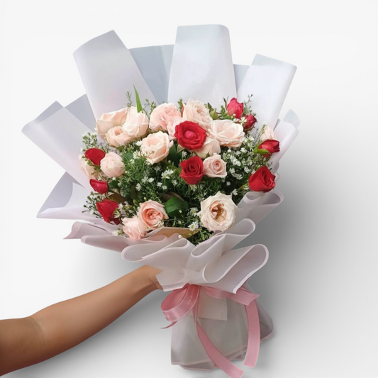 The Flowercart Blushing Harmony bouquet features 24 pink and red roses, elegantly wrapped in white paper with a pink ribbon. Perfect for romantic occasions.