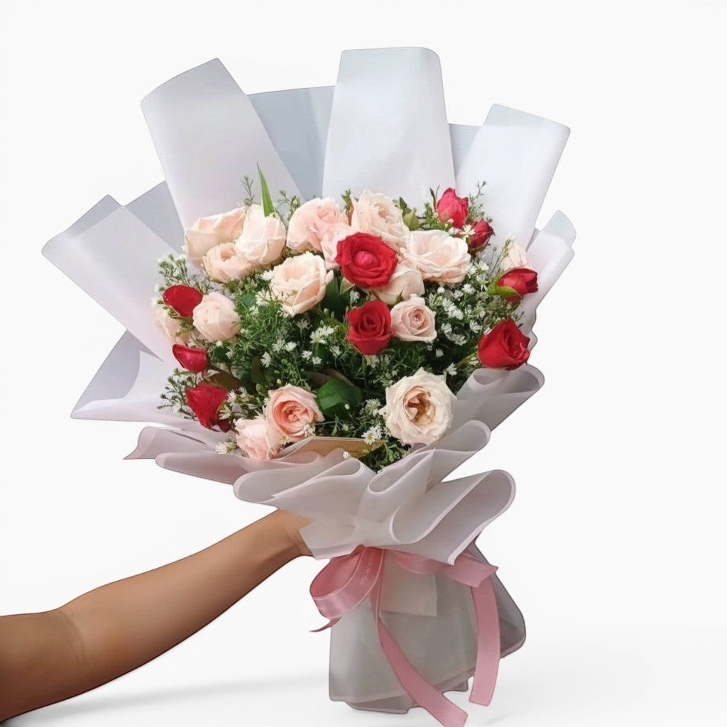 The Flowercart Blushing Harmony bouquet features 24 pink and red roses, elegantly wrapped in white paper with a pink ribbon. Perfect for romantic occasions.