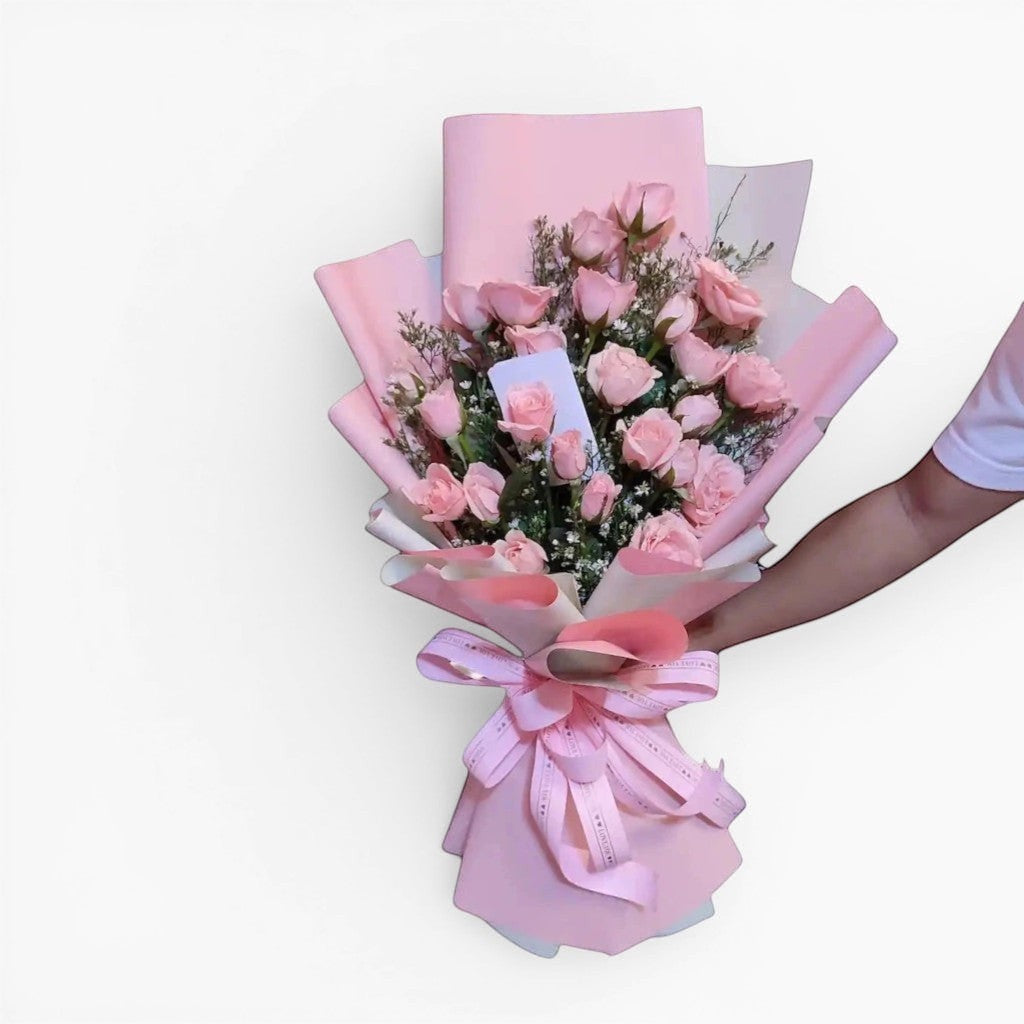The Flowercart Pink Whisper bouquet features 24 premium pink roses and baby's breath, elegantly wrapped in pink paper with a matching ribbon. Ideal for romantic occasions.