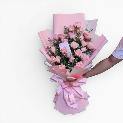 The Flowercart Pink Whisper bouquet features 24 premium pink roses and baby's breath, elegantly wrapped in pink paper with a matching ribbon. Ideal for romantic occasions.
