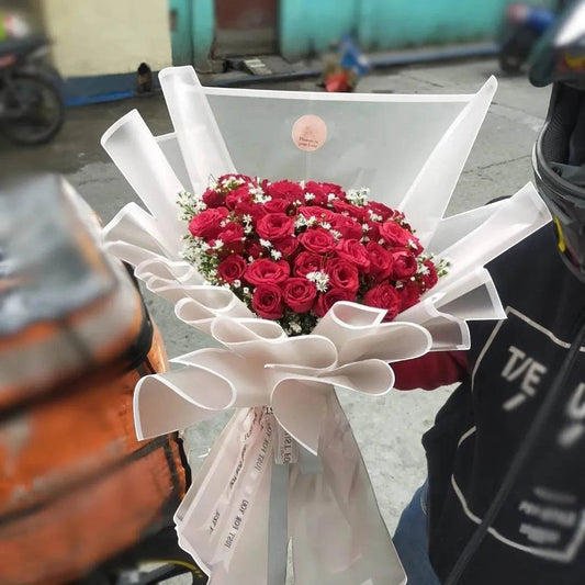 The Flowercart's Scarlet bouquet features 50 vibrant red roses with delicate baby's breath, elegantly wrapped in white paper.