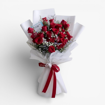 The Flowercart's Fallin For You bouquet features 24 vibrant red roses with baby's breath and misty blue fillers, elegantly wrapped in white paper with a red ribbon.