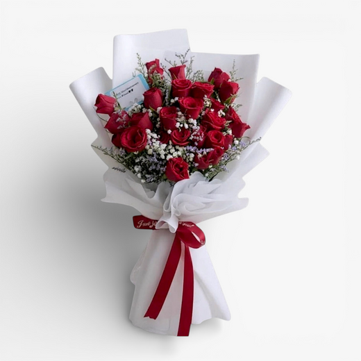 The Flowercart's Fallin For You bouquet features 24 vibrant red roses with baby's breath and misty blue fillers, elegantly wrapped in white paper with a red ribbon.