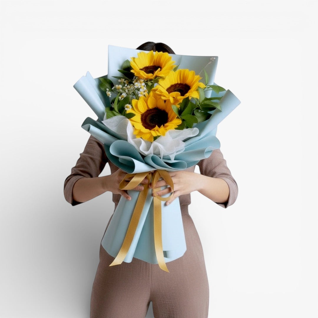 The Flowercart Sunshine Delight bouquet features three large sunflowers with complementary yellow and white blooms, wrapped in light blue paper and tied with a gold ribbon.