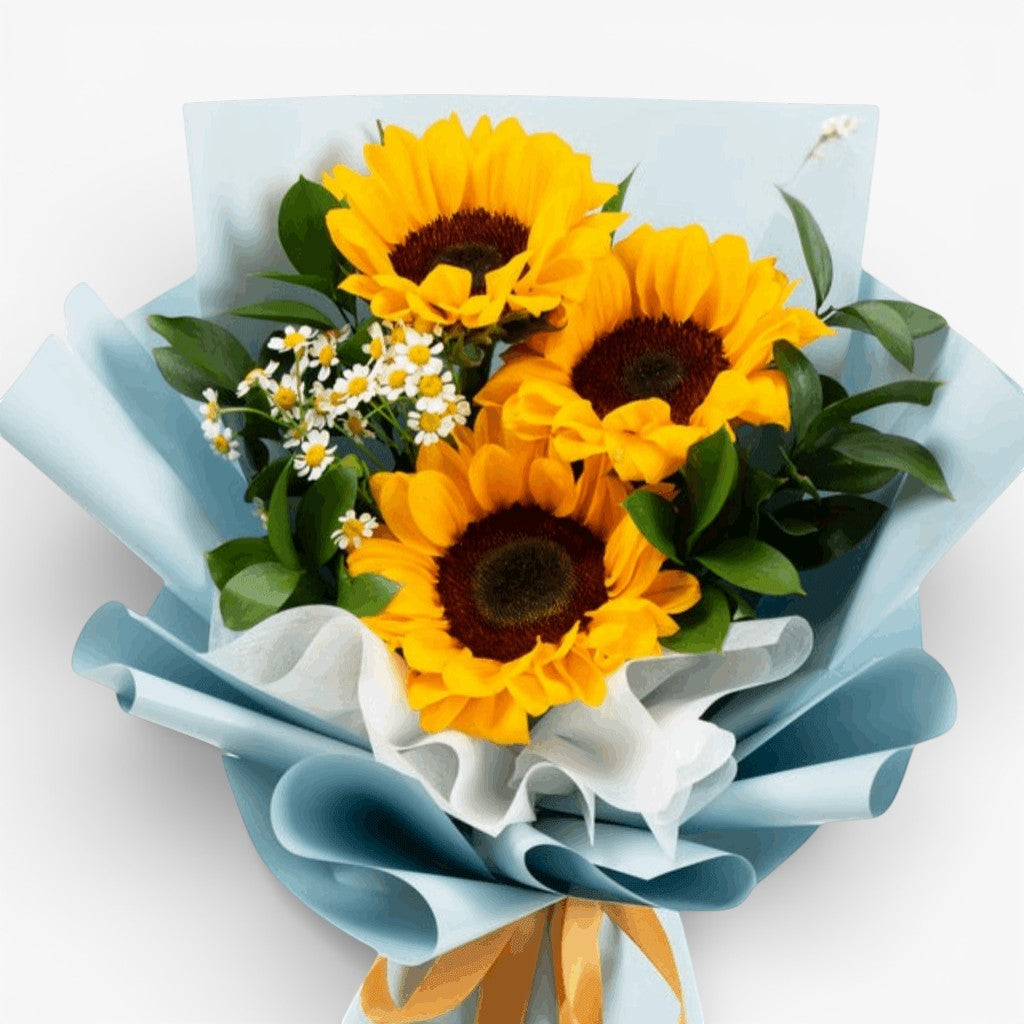The Flowercart Sunshine Delight bouquet features three large sunflowers with complementary yellow and white blooms, wrapped in light blue paper.