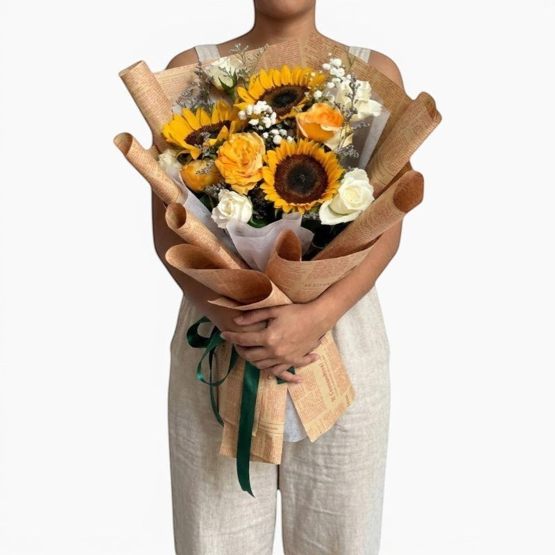 The Flowercart Sunny Serenade bouquet: sunflowers, yellow and white roses, baby's breath, and misty blue fillers in brown paper wrap.