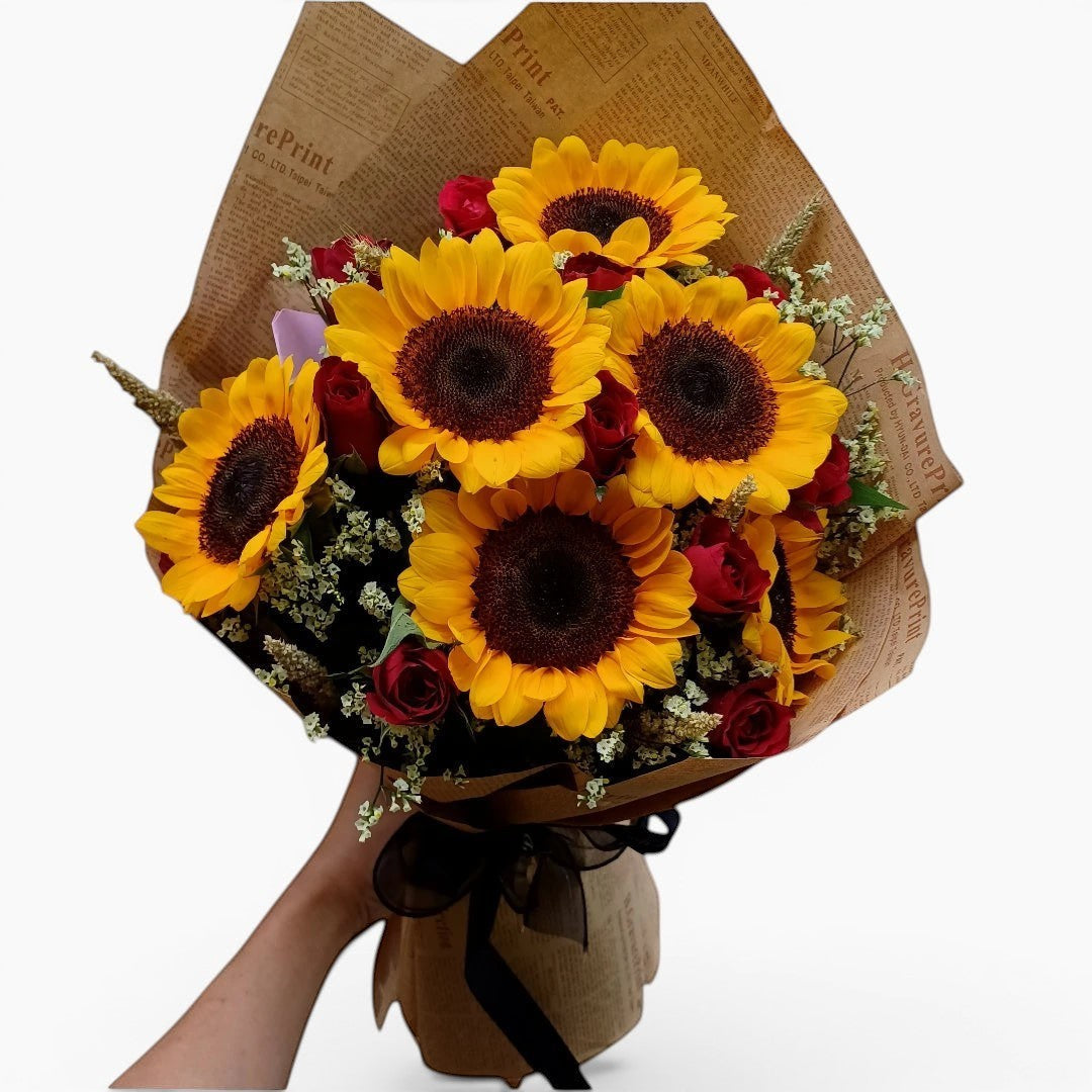 The Flowercart's Sunny Blooms bouquet features 6 golden sunflowers and 12 red roses with baby's breath, wrapped in brown paper.