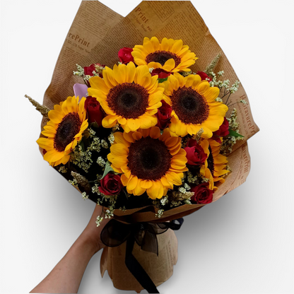 The Flowercart's Sunny Blooms bouquet features 6 golden sunflowers and 12 red roses with baby's breath, wrapped in brown paper.