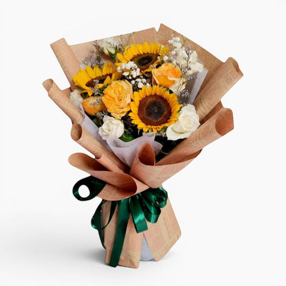 The Flowercart Sunny Serenade bouquet features sunflowers, white and yellow roses, baby's breath, and misty blue fillers, wrapped in brown paper and tied with a green ribbon.