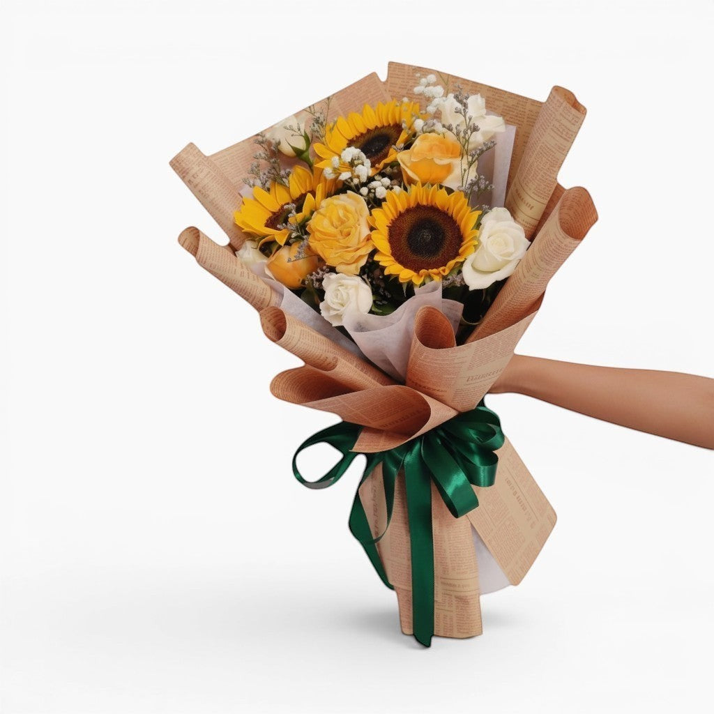 The Flowercart Sunny Serenade bouquet features sunflowers, yellow roses, white roses, and baby's breath, wrapped in brown paper with a green ribbon.