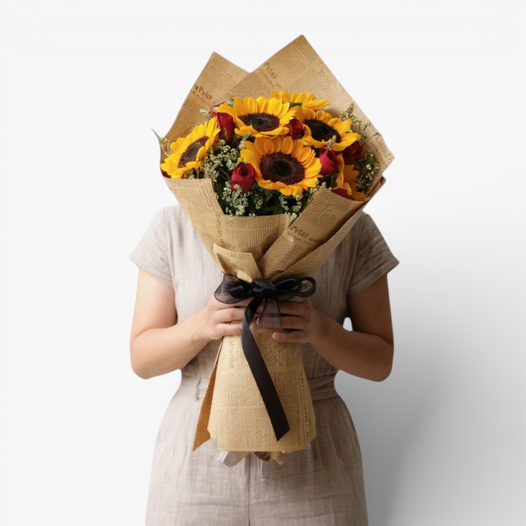 The Flowercart's Sunny Blooms bouquet: 6 golden sunflowers, 12 red roses, and baby's breath, wrapped in newspaper-style paper with a black ribbon.